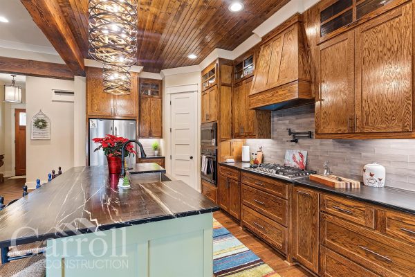 a different angle of a custom kitchen with stained oak cabinets, pine ceilings and modern chandeliers. Undercounter lighting displaying a linear pattern backsplash near a black soapstone.