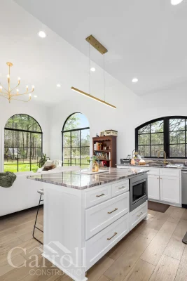 Angled view of kitchen island with linear brushed brass pendant and large brass chandelier over living room